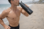 Load image into Gallery viewer, Man drinking from a black 'bamba' water bottle on a beach.
