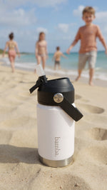 Load image into Gallery viewer, White bamba water bottle with black lid and straw on a sandy beach with people in the background.
