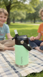 Load image into Gallery viewer, Green bamba kids water bottle with black lid on a checkered blanket in a park setting
